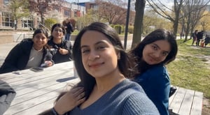 A group of four friends&nbsp;sitting&nbsp;around a&nbsp;table outside on a sunny day. The girl&nbsp;in the front is&nbsp;taking&nbsp;a selfie while smiling at the camera. The three women behind her are smiling too&nbsp;. The background includes the&nbsp;university campus with trees, open sky&nbsp;, and a building.