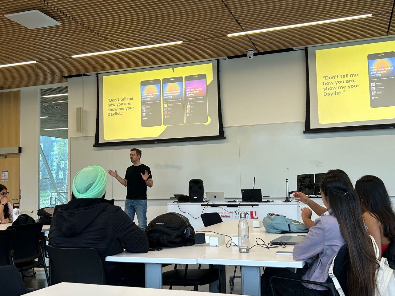Ivan Peher, country manager at Spotify, presenting for students at Marketing for Search and Social Media class. Students sitting in front of them behind white tables paying attention to his slides on projectors. white board is clean and laptops and water bottles are on the tables.