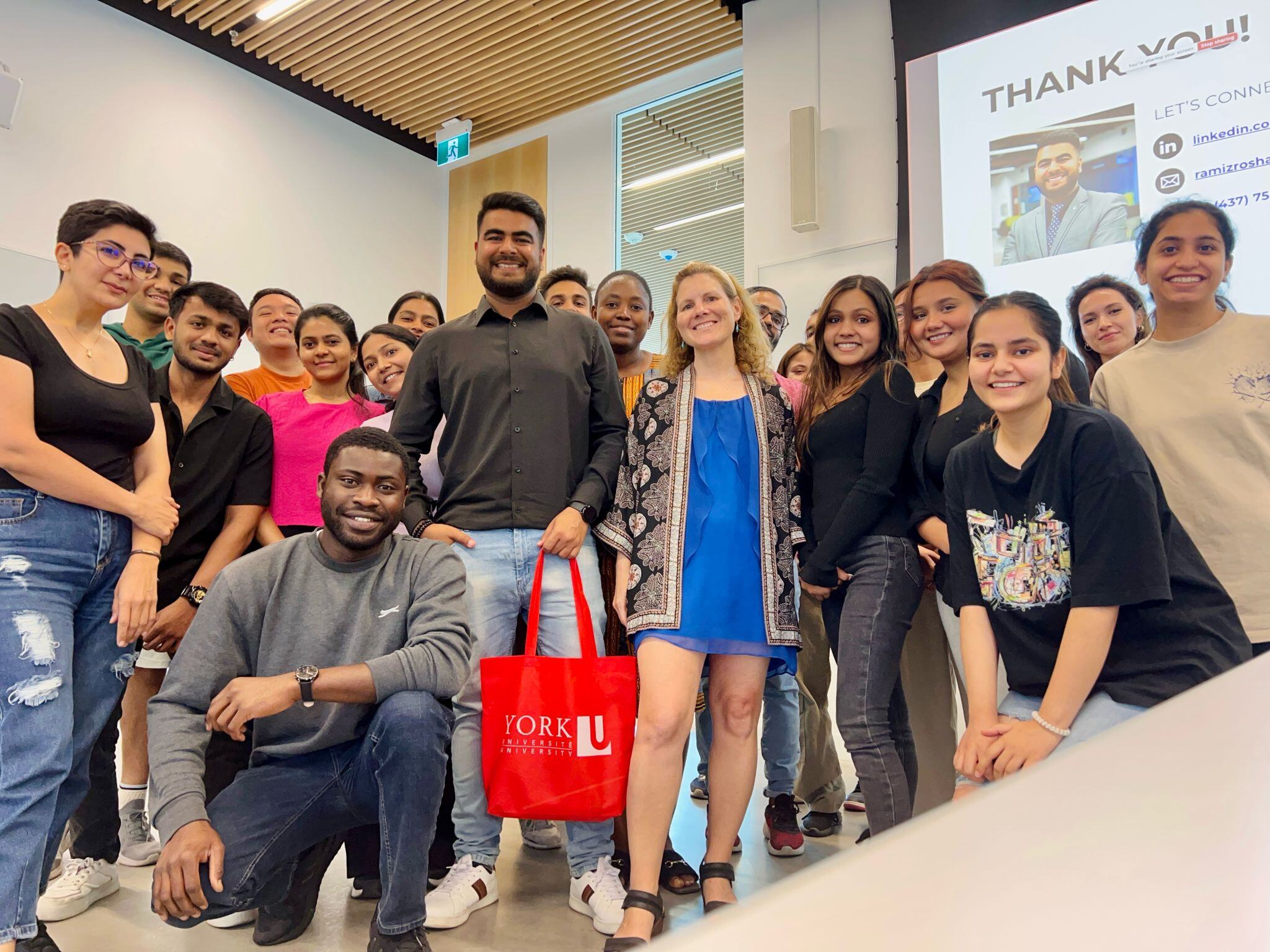 A group photo of students and their professor in a classroom with a guest speaker holding a red York University promotional bag, all smiling and looking at the camera.