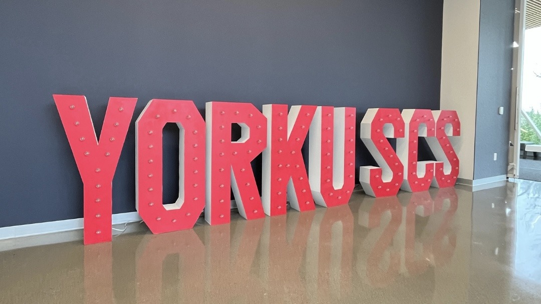 York University School of Continuing Studies signage in the campus hallway in big red bold letters that read YORKU SCS, lights up in the dark. Red signage is placed against black wall on the tiled floor.