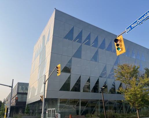 Exterior view of York University's School of Continuing Studies building, showcasing its modern glass facade and angular design, with a clear blue sky above. and another building adjacent to it. A traffic light which is green is visible along with a tree. 
