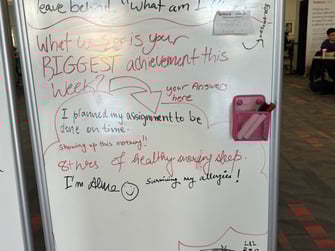 A white board with a question written on it with a red marker. The question is What was or is your biggest achievement this week with the word biggest written in all caps. There are 5 different answers written in different handwritings. Ans1-I planned my assignment to be done on time written with black marker. Ans2 and 3- Showing up this morning! and 8hrs of healthy snoring sleep written with red marker Ans 4and 5- I am alive (smiley emoji) and Surviving my allergies! written with a black marker 