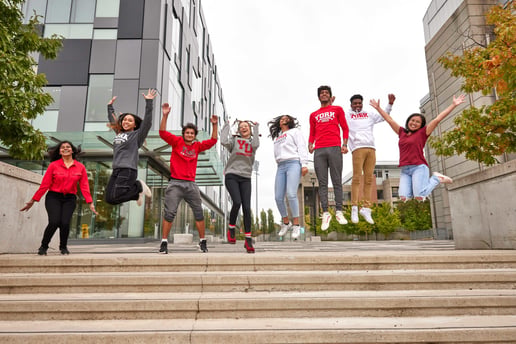 The image shows a group of seven young adults, university students, mid-jump on an outdoor staircase in front of a modern campus building. They are all smiling and appear to be enjoying themselves. Most of them are wearing casual attire, including sweatshirts and T-shirts with "York University" printed on them, indicating they are students of York University. The background includes trees and contemporary architectural structures, suggesting a university campus setting.
