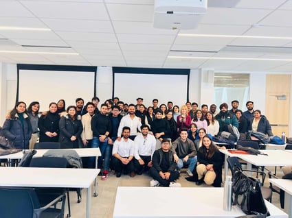 A group of students posing in a classroom with tables, chairs and projector.