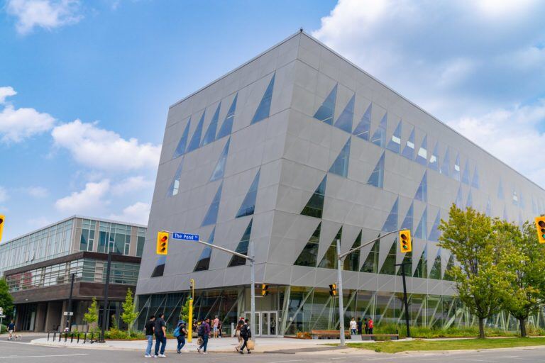 Alt: York University School of Continuing Studies Building a modern, geometrically designed building with glass panels under a clear blue sky. The scene is animated with pedestrians crossing the street in front of this architectural marvel.