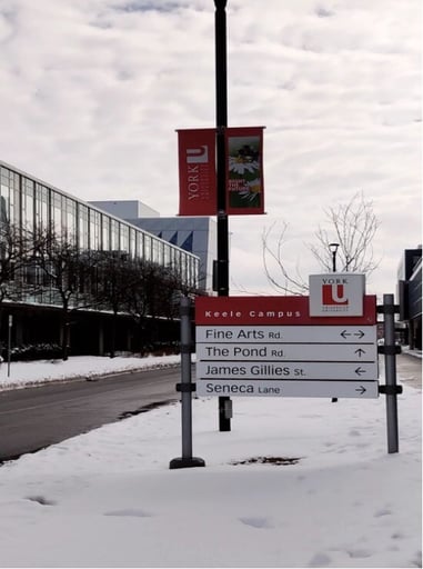 Snow-covered campus on a winter morning with a signboard pointing to buildings, under a cloudy sky