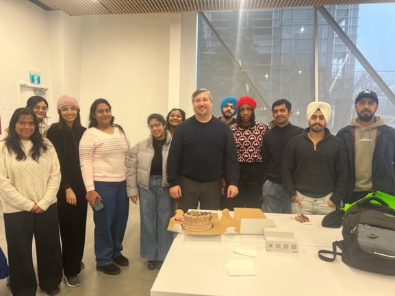 tudents celebrating a professor's birthday, standing in front of a cake and smiling