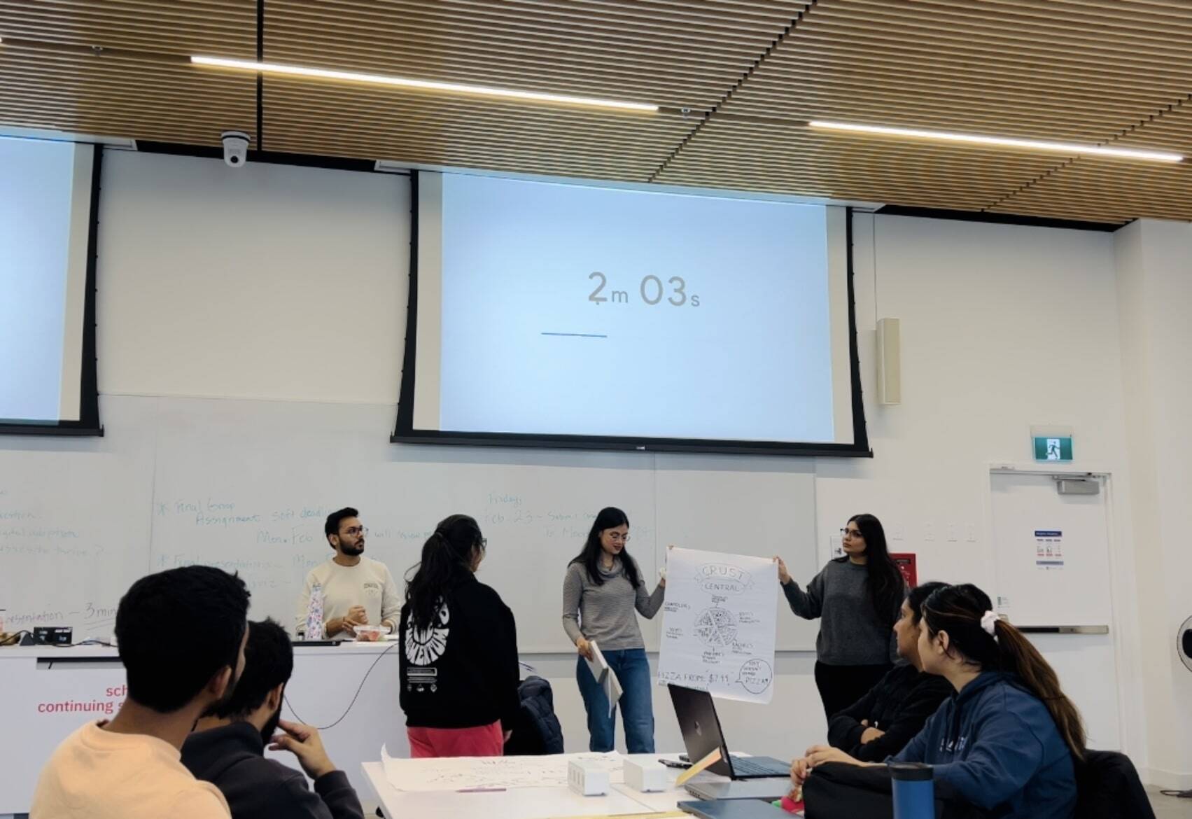 A group of four digital marketing post-graduate students presenting a marketing pitch in their classroom while the timer goes on and the students are attentively listening.