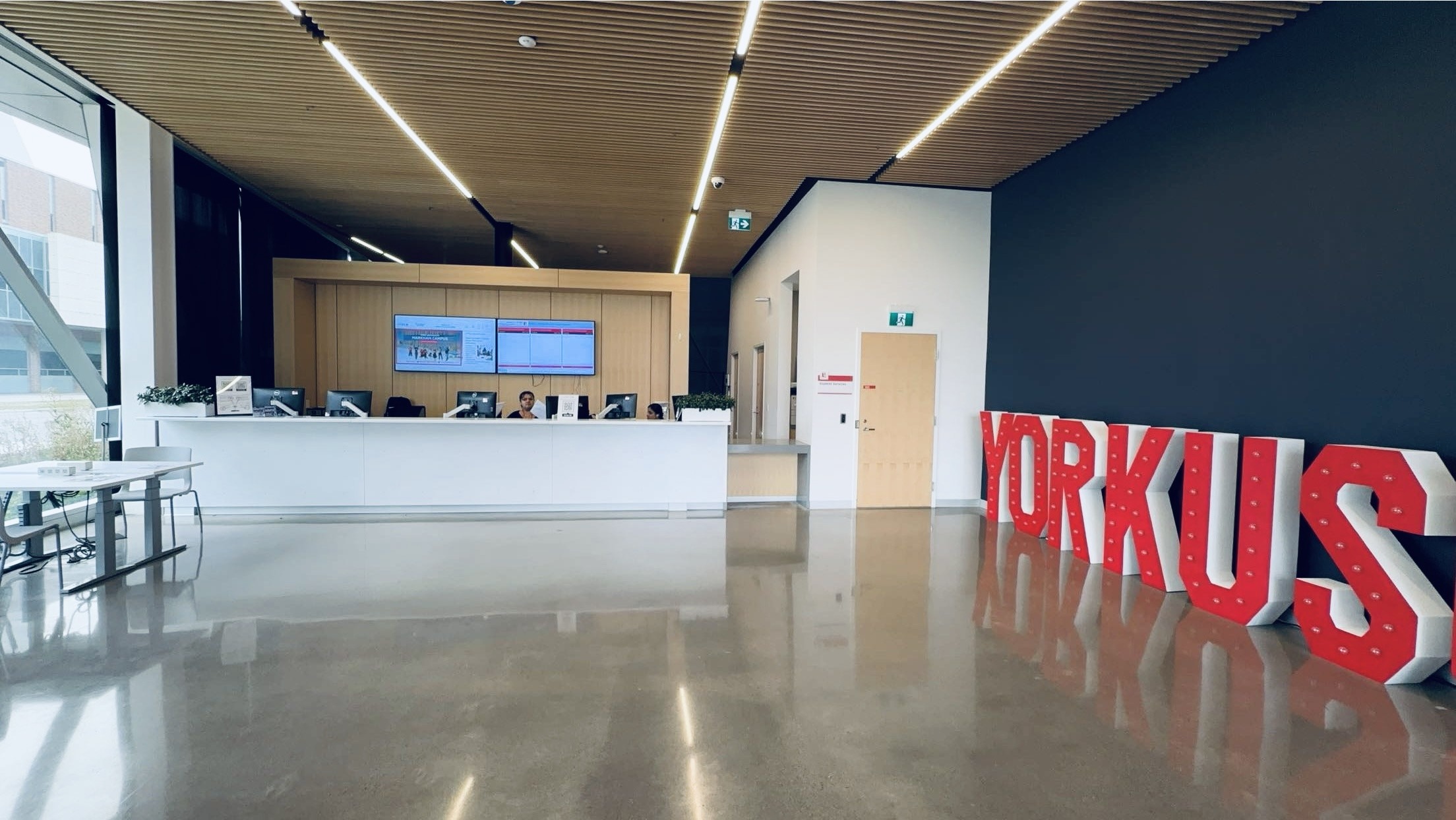 A sleek reception area with a white desk, overhead screens, and large red 