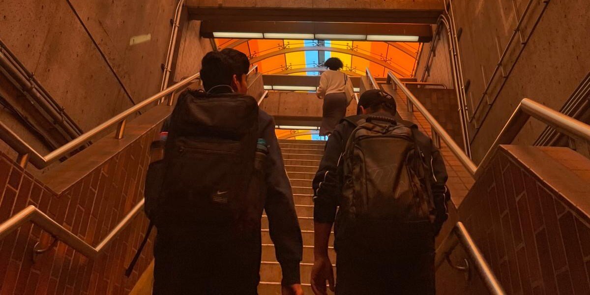 Two young males climbing up a flight of stairs side by side with backpacks, a combination of red and yellow hue as the sun hits the ceiling, and a female at the top of the stairs in the middle of the frame