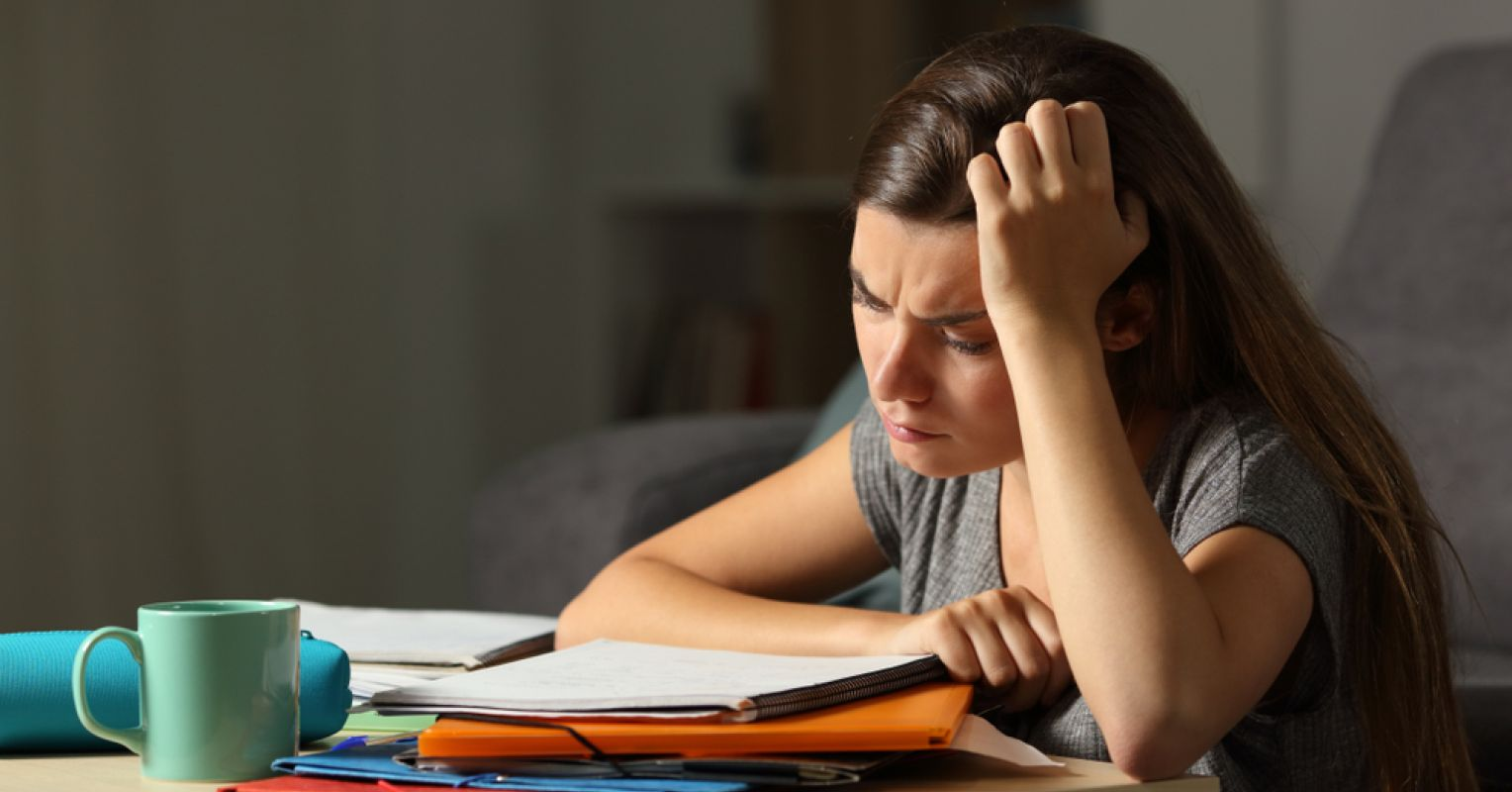 A university student is looking over her notebook, seemingly worried about not completing her work on time. There’s a coffee cup on the table, which is evidence that she may be pulling an all-nighter to complete her work on time.