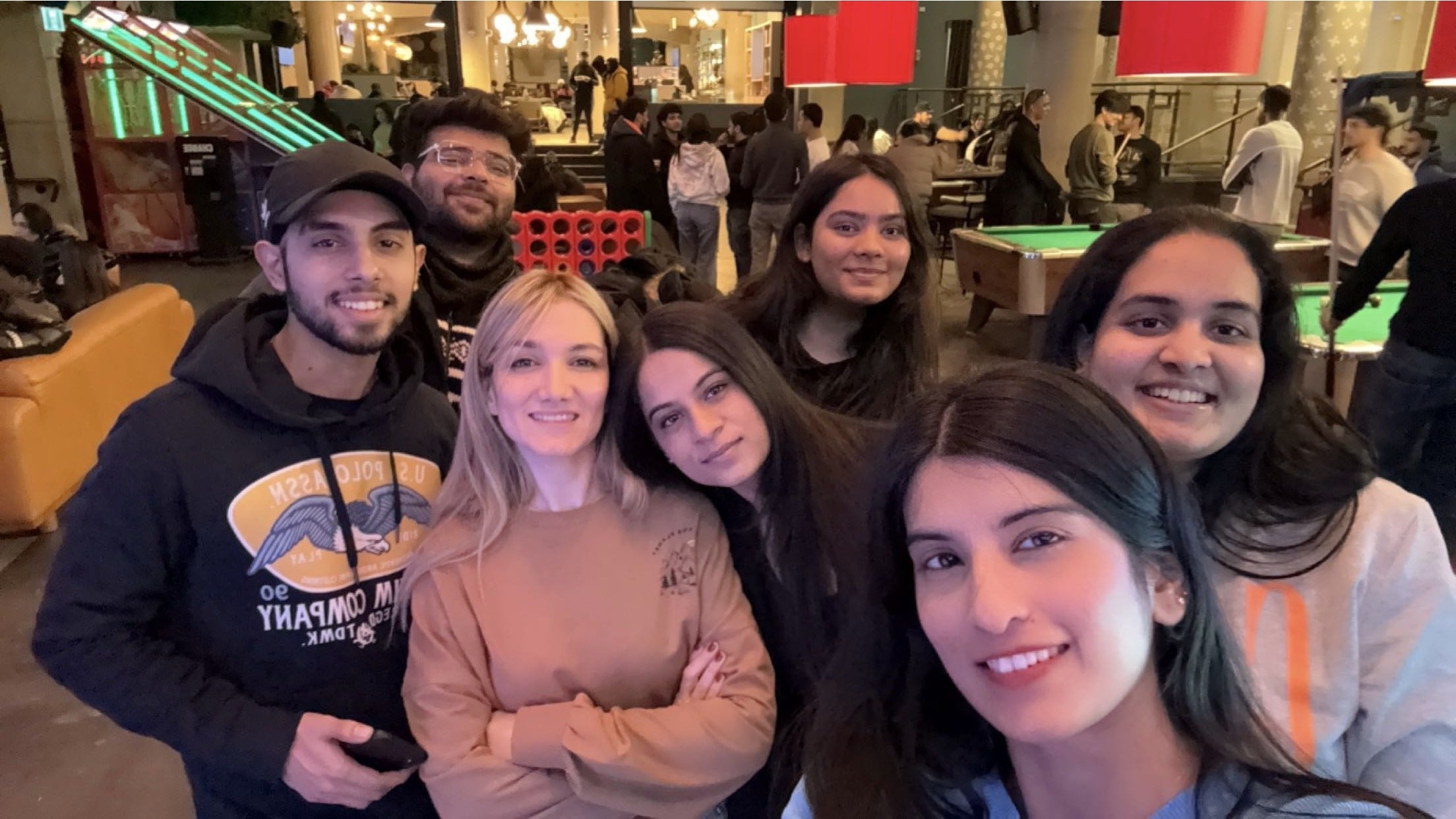 A group of seven people smiling and posing for a selfie in a lively indoor setting with other people in the background, including pool tables and a giant Connect Four game.